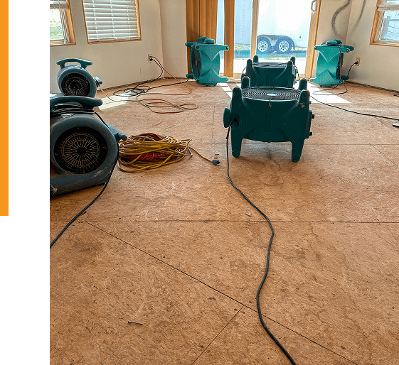 Group 9 (4) Room with plywood floor, several industrial air movers, tangled cords, and sunlight coming through windows and a glass door, suggesting ongoing water damage restoration or drying process.