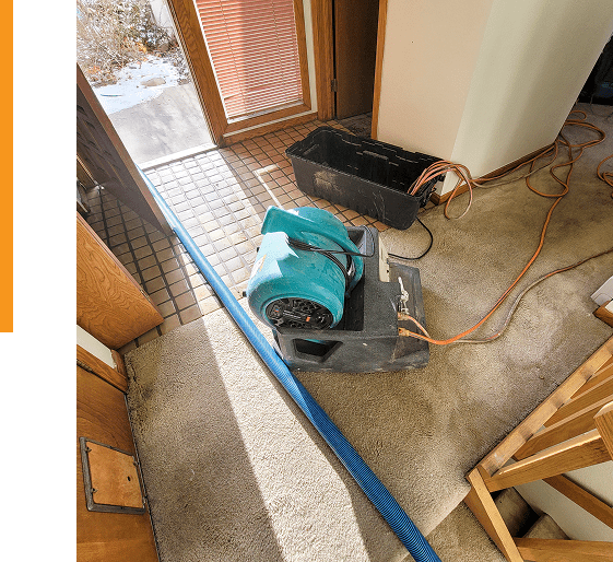 Group 9 (5) A blue air mover and a black plastic bin sit on beige carpet near a tiled entryway, with extension cords and a blue hose running across the floor.